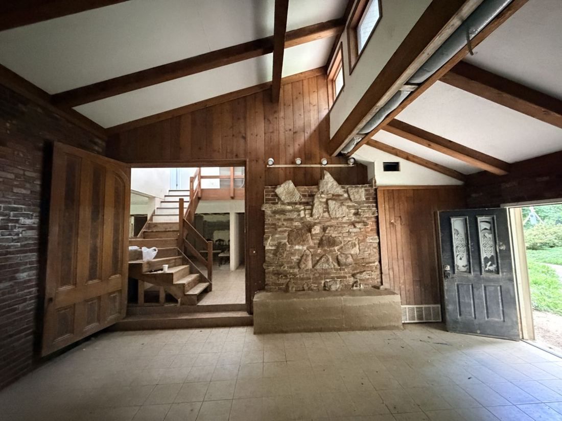 Great room with exposed beams, a stone fireplace and an open staircase before renovation